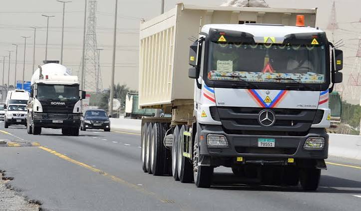 A white dump truck and a cement mixer truck driving on a multi-lane highway in the UAE, with cars, street lights and power pylons behind them.